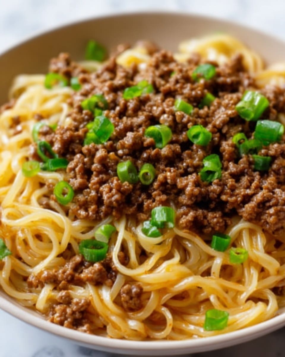 The image shows a close-up of a bowl of noodles topped with cooked ground beef and chopped green onions. The noodles are yellowish and look soft with a slightly glossy texture, forming the bottom layer filling most of the bowl. On top, the ground beef is scattered evenly across the noodles, appearing brown and crumbly. Small pieces of bright green chopped onions are sprinkled over the beef, adding color contrast. The bowl is white and sits on a white marbled surface. Photo taken with an iphone --ar 4:5 --v 7