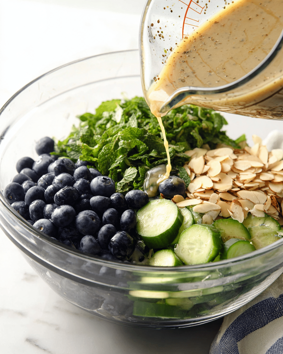 A clear glass bowl shows four main layers of ingredients: bright green chopped mint leaves on the upper left, dark blue whole blueberries next to them on the right, medium green cucumber slices with white flesh below the mint, and light brown thin sliced almonds below the blueberries and cucumber, all gathered closely together. A glass measuring cup is tilted above the bowl, pouring a light tan dressing with visible brown specks over the ingredients. The bowl sits on a white marbled surface with part of a folded white and blue cloth visible in the lower right corner. photo taken with an iphone --ar 4:5 --v 7