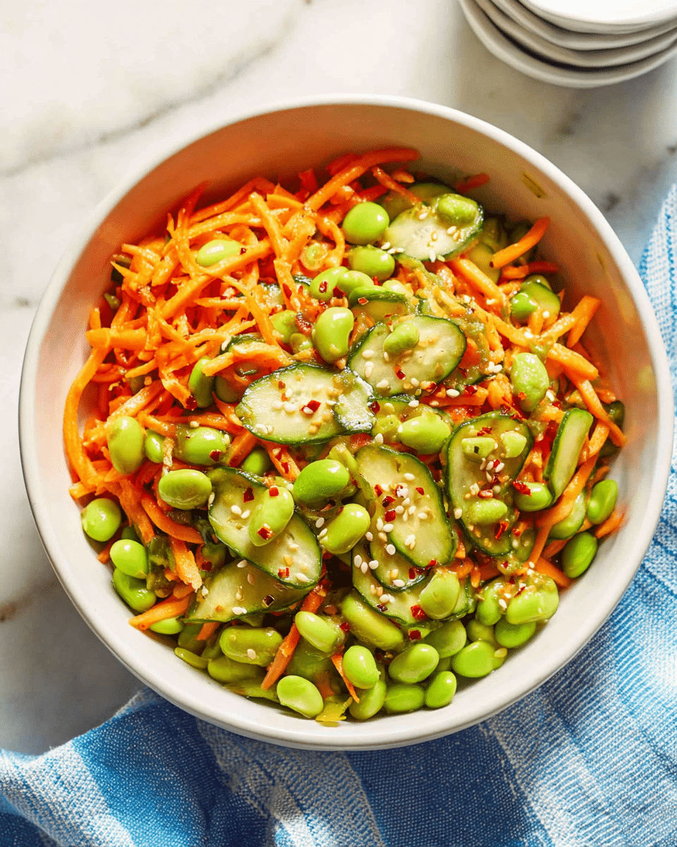 A white bowl filled with a fresh salad made of three main layers: bright green edamame beans, thin, bright orange shredded carrots, and light green cucumber slices with dark green edges. The salad is mixed together and topped with small white sesame seeds and red chili flakes. The bowl sits on a white marbled surface with a blue and white striped cloth nearby, and there are white stacked cups in the background. Photo taken with an iphone --ar 4:5 --v 7