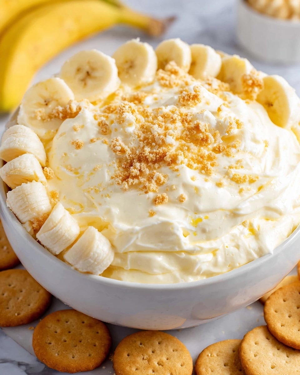 A white bowl filled with a thick, creamy light yellow mixture that has a fluffy texture, sprinkled with small crumbled golden brown pieces on top. This creamy layer is surrounded by a ring of evenly sliced banana pieces, each slice showing a soft yellow color with pale edges and a central dot pattern. The bowl rests on a white marbled surface, with small round golden brown cookies placed around its base. Photo taken with an iphone --ar 4:5 --v 7