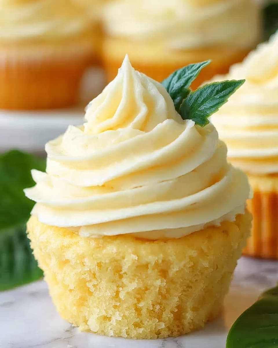 The image shows a close-up of a vanilla cupcake with two main layers. The bottom layer is a golden yellow, soft cake with a slight crumbly texture. On top, there is a thick swirl of creamy white frosting, piped in a smooth spiral with a pointed peak at the center. A small green leaf decoration sticks out from the frosting near the peak, adding a fresh contrast to the pale colors. The background has a white marbled texture with more cupcakes slightly blurred behind. photo taken with an iphone --ar 4:5 --v 7