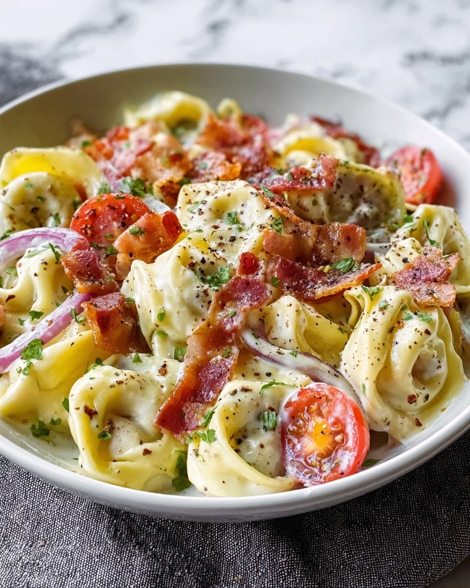 A close-up view of a white bowl filled with creamy tortellini pasta, showing about two layers of tightly folded yellow pasta pieces coated in white sauce. Interspersed among the pasta are bright red cherry tomato halves, crispy brown bacon bits, and thin slices of light purple onion, all sprinkled with freshly chopped green herbs. The dish is topped with a light dusting of black pepper and some grated white cheese. The bowl sits on a textured light gray cloth over a white marbled surface. photo taken with an iphone --ar 4:5 --v 7
