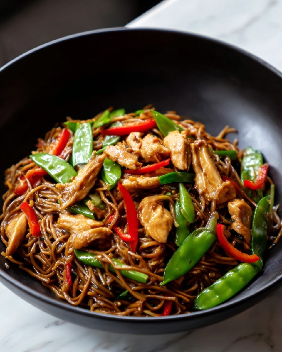 A black bowl filled with a noodle dish featuring multiple layers of thin brown noodles mixed with small pieces of cooked chicken. Bright green sugar snap peas and thin slices of red chili peppers are scattered throughout, adding pops of color. The noodles and vegetables have a glossy, slightly oily texture. The bowl sits on a white marbled surface. Photo taken with an iphone --ar 4:5 --v 7