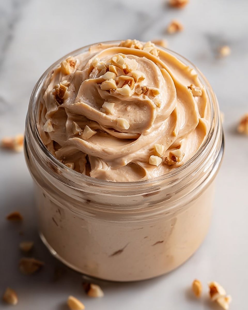 A clear glass jar filled with smooth, creamy peanut butter mousse that is light tan in color with soft, swirling peaks covering the entire jar. The top layer is decorated with chopped peanuts scattered unevenly, adding a bit of texture and off-white color contrast. The jar is placed on a white marbled surface with some peanut pieces around it, and a blurred similar jar is visible in the background. photo taken with an iphone --ar 4:5 --v 7