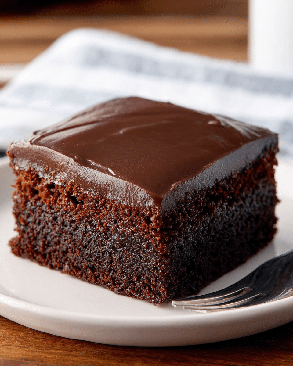 A single square piece of rich chocolate cake sits on a white round plate. The cake has two layers: the bottom layer is a dark, dense chocolate base, while the top layer is a lighter, moist chocolate sponge. The top of the cake is covered with a smooth, thick layer of glossy dark chocolate frosting. The plate is placed on a wooden surface with a blurred white marbled texture background, and a silver spoon is partially visible at the edge. photo taken with an iphone --ar 4:5 --v 7