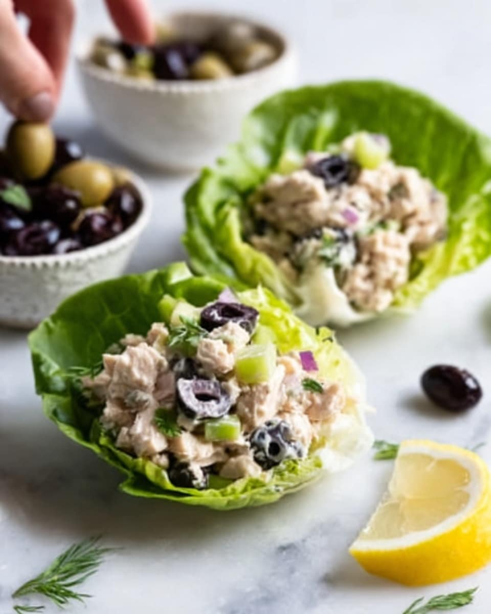 The image shows two lettuce cups filled with a creamy salad that has visible chunks of chicken, diced celery, and blueberries mixed in. The lettuce leaves are fresh and bright green, forming natural bowls that hold the colorful salad. On the white marbled surface around the cups, there are a few scattered blueberries, a sprig of rosemary, and a lemon wedge, adding a fresh and vibrant feel to the scene. In the background, a small white bowl filled with blueberries is partially visible. The photo was taken with an iphone --ar 4:5 --v 7