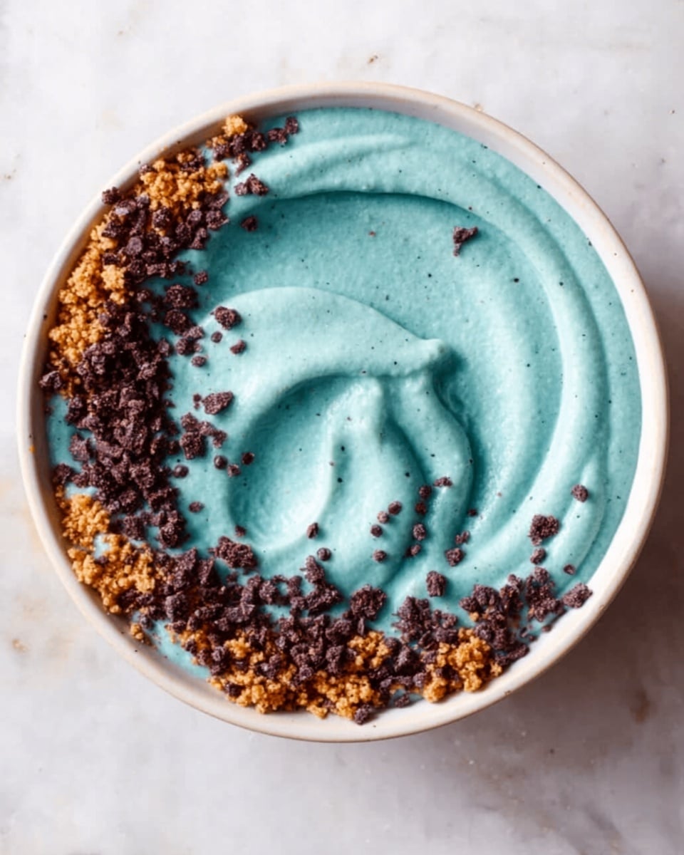 A white bowl filled with a smooth, thick blue-green creamy base that occupies the center, swirled gently in the middle to create a small peak. Around the top edge of the bowl, there is a ring of crumbly light brown pieces mixed with small dark brown crunchy bits, giving a textured contrast to the creamy center. The bowl is placed on a white marbled surface, and some of the crumbly topping is scattered loosely outside the bowl near the edges. photo taken with an iphone --ar 4:5 --v 7