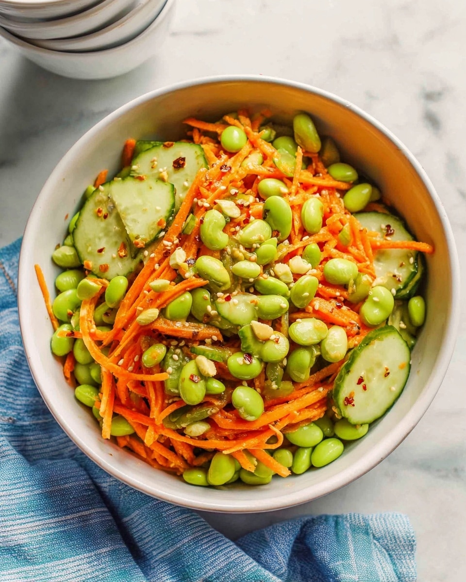 A white bowl filled with a colorful salad sits on a white marbled surface. The salad has three main layers: thin orange carrot sticks, bright green edamame beans, and slices of pale green cucumber. The vegetables are mixed evenly, with a glossy sauce that adds shine and some red chili flakes scattered throughout. White sesame seeds are sprinkled on top, adding texture and contrast to the fresh ingredients. A striped cloth napkin and a stack of white bowls with gold rims are partially visible in the background. photo taken with an iphone --ar 4:5 --v 7